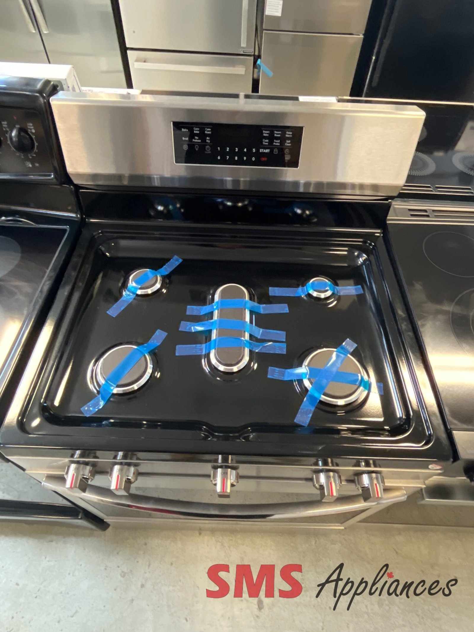 Open-Box-Stoves-Ranges-GCRG3060BFE-3-scaled-1.jpg Stainless steel gas range with control panel above a black oven, blue tape on burners, showroom floor.