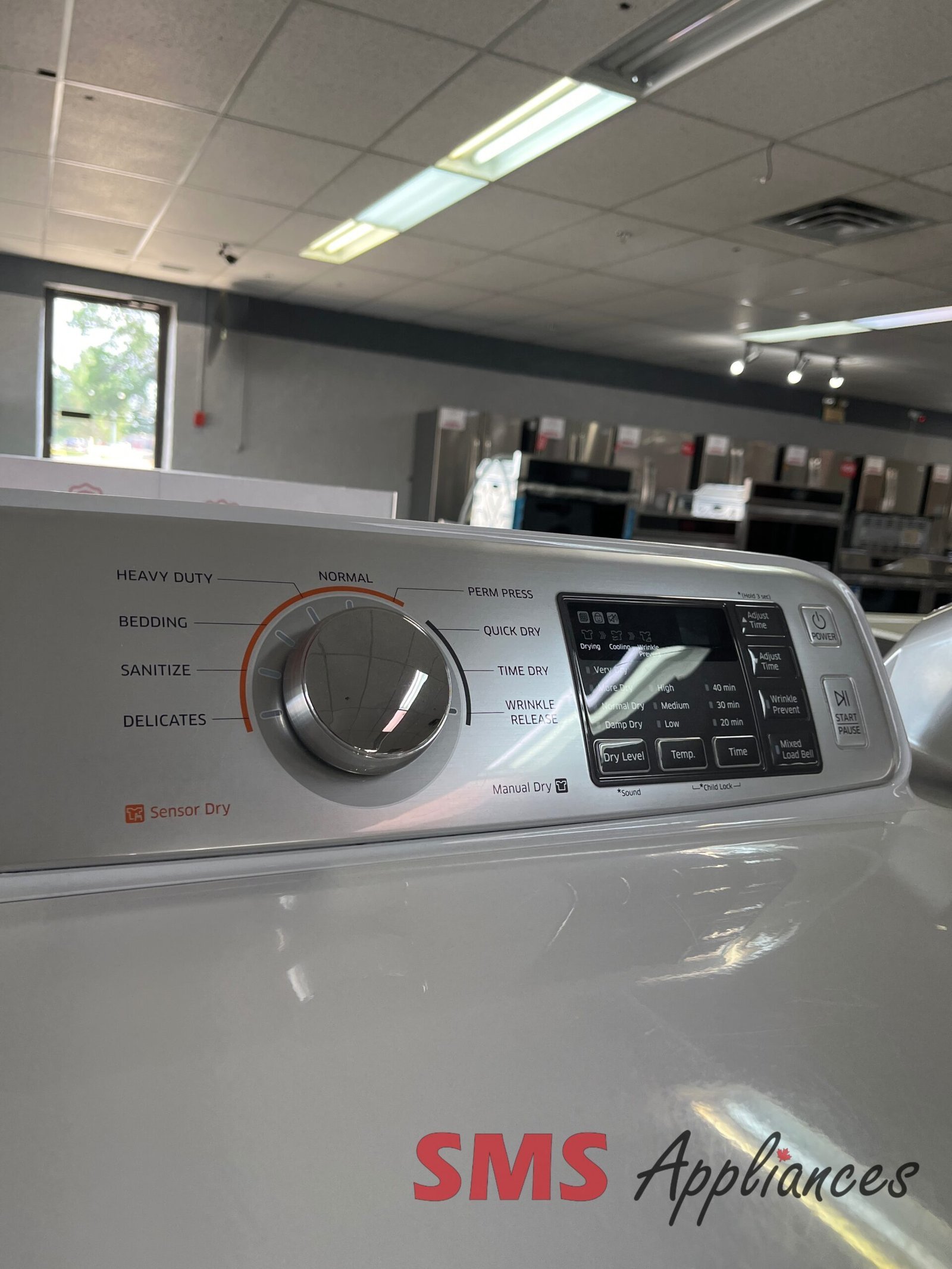 Refurbished-Dryers-DV45H7000EW-4-scaled-1.jpg Front-loading washing machine control panel with a large dial and digital display in an appliance showroom; 'SMS Appliances' logo visible on the machine side.