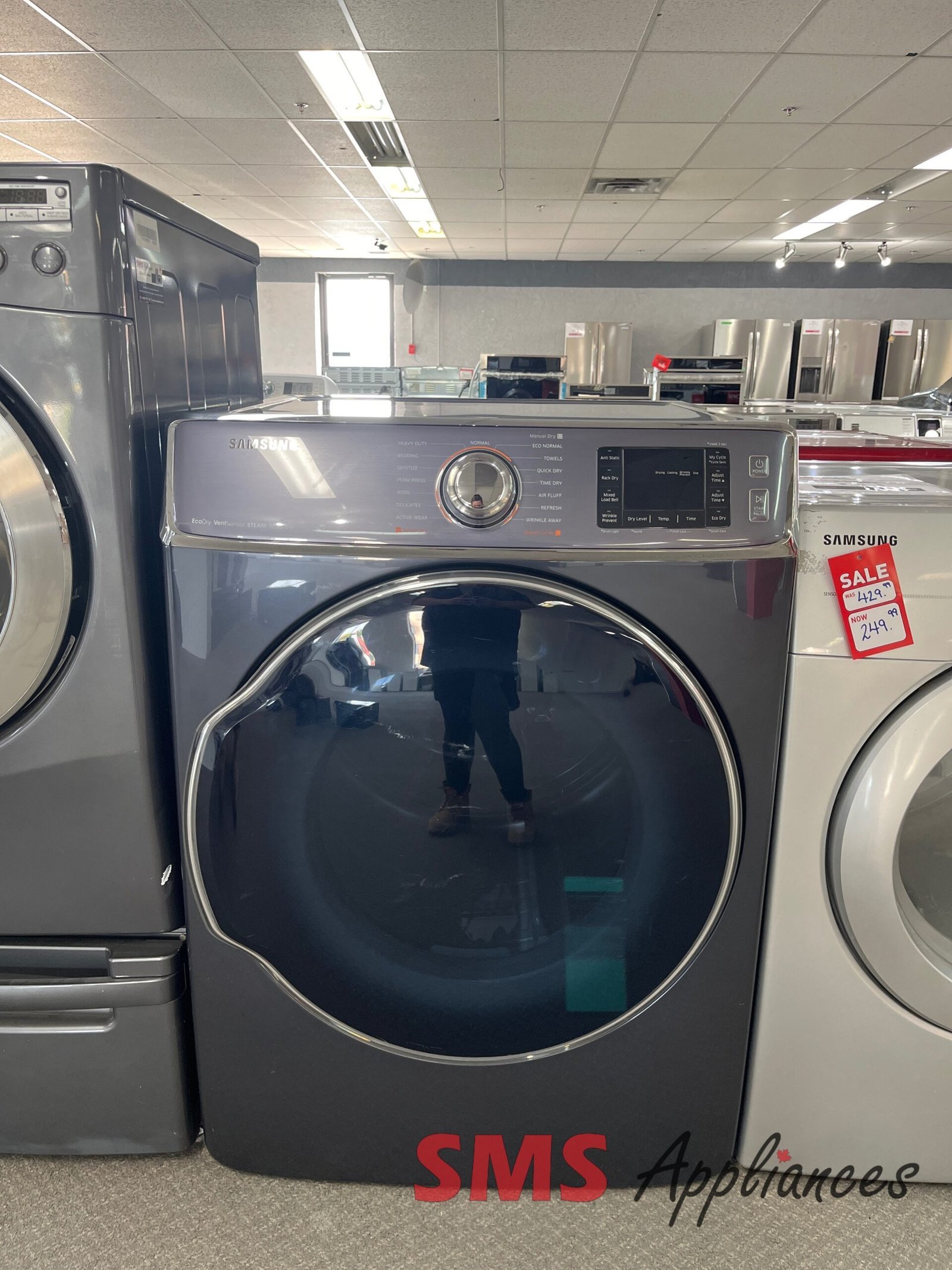 Refurbished-Dryers-DV56H9100EG-1-scaled-1.jpg Front-loading Samsung washing machine on a showroom floor with other stainless appliances in the background and an 'SMS Appliances' sign at the base.